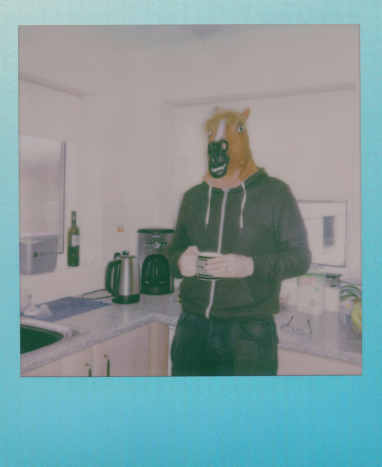 A person wearing a horse mask stands in a kitchen holding a mug, captured on instant film.